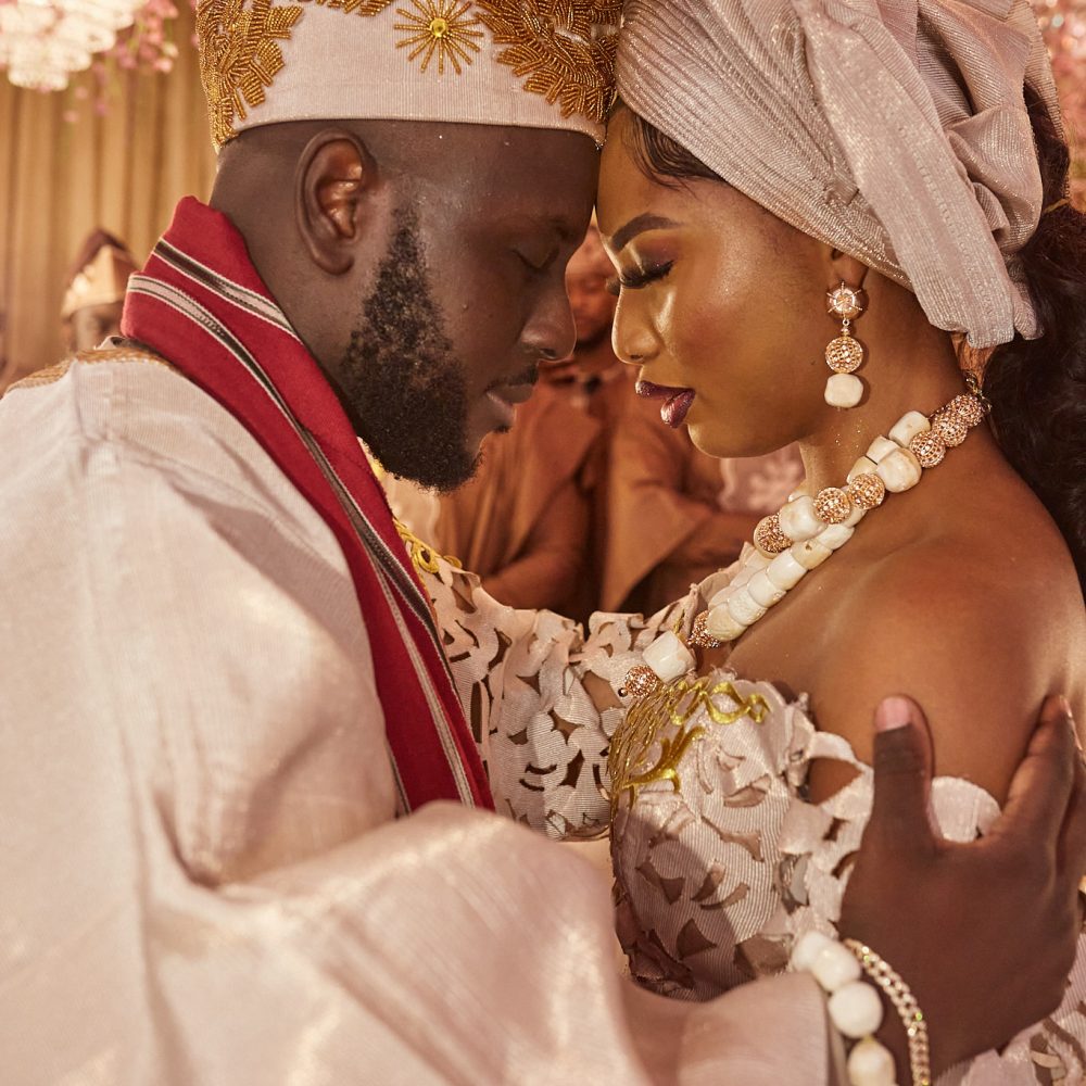 Bride and groom forehead to forehead in cream traditional attire and coral beads