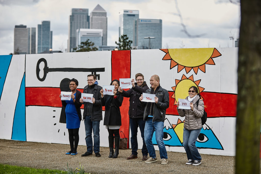 Friends holding up proposal signs in front of a colourful mural with the London skyline behind