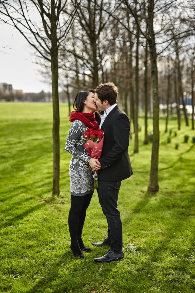 Newly engaged couple kissing under bare trees, the woman holding red roses