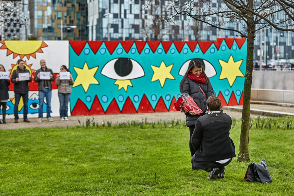 Man on one knee proposing in front of a mural as friends hold up signs