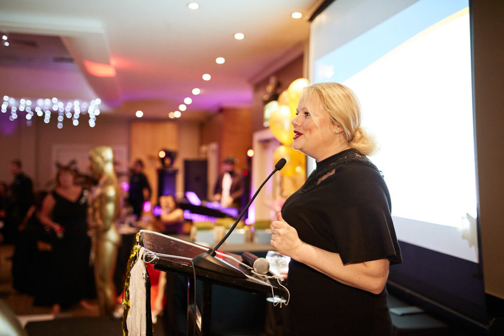 Host speaking at the lectern beside the presentation screen