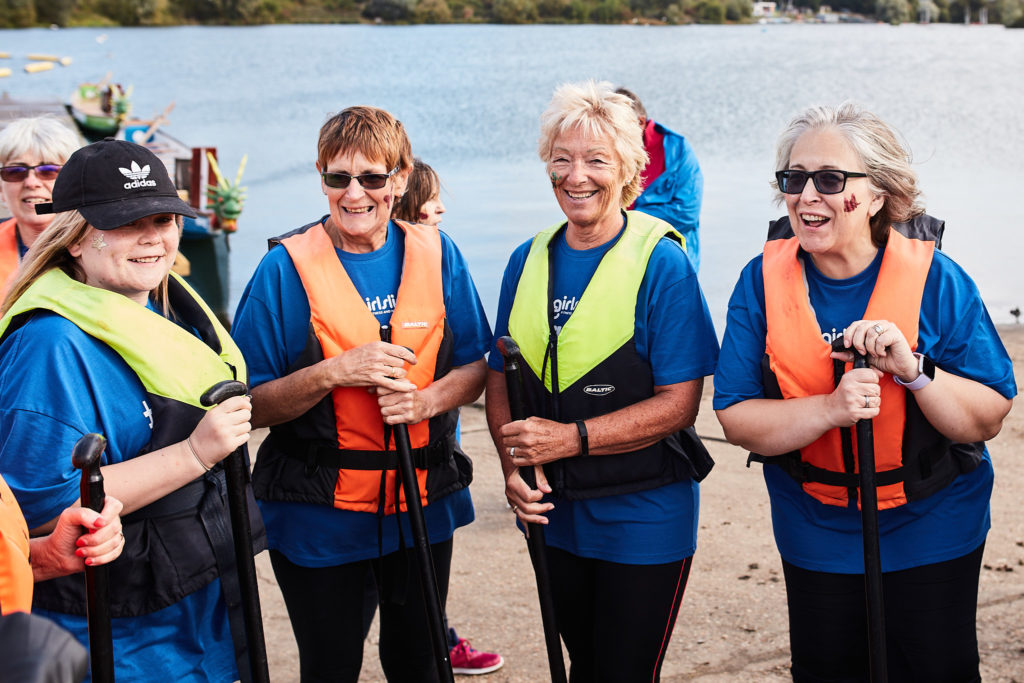 Four women in blue team shirts and life jackets smiling beside the lake