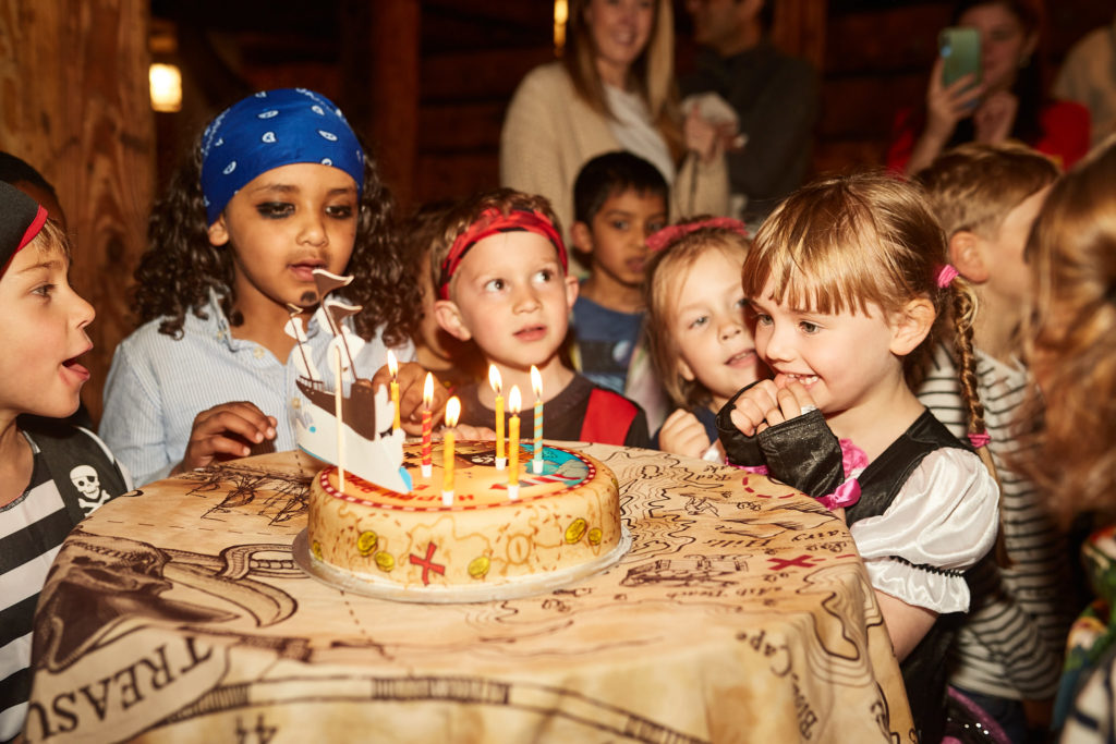 Children gathered round a treasure chest birthday cake with lit candles