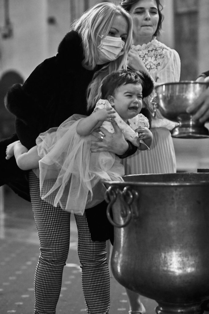 Black and white photo of a crying toddler being lifted beside the baptism font