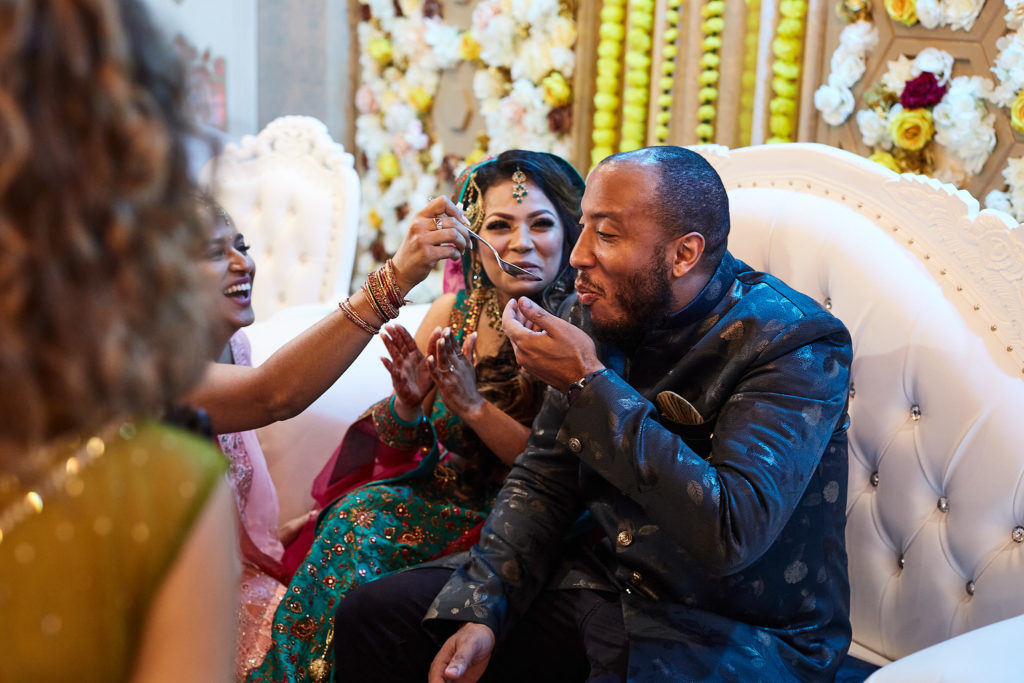 Bride-to-be laughing with the groom on a white sofa as a guest reaches towards them