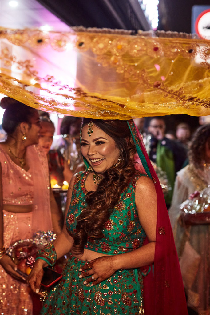 Bride-to-be smiling in a green lehenga beneath a gold canopy during her mehndi procession