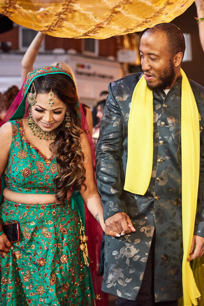 Bride in a green lehenga and groom in a yellow scarf walking hand in hand beneath the canopy