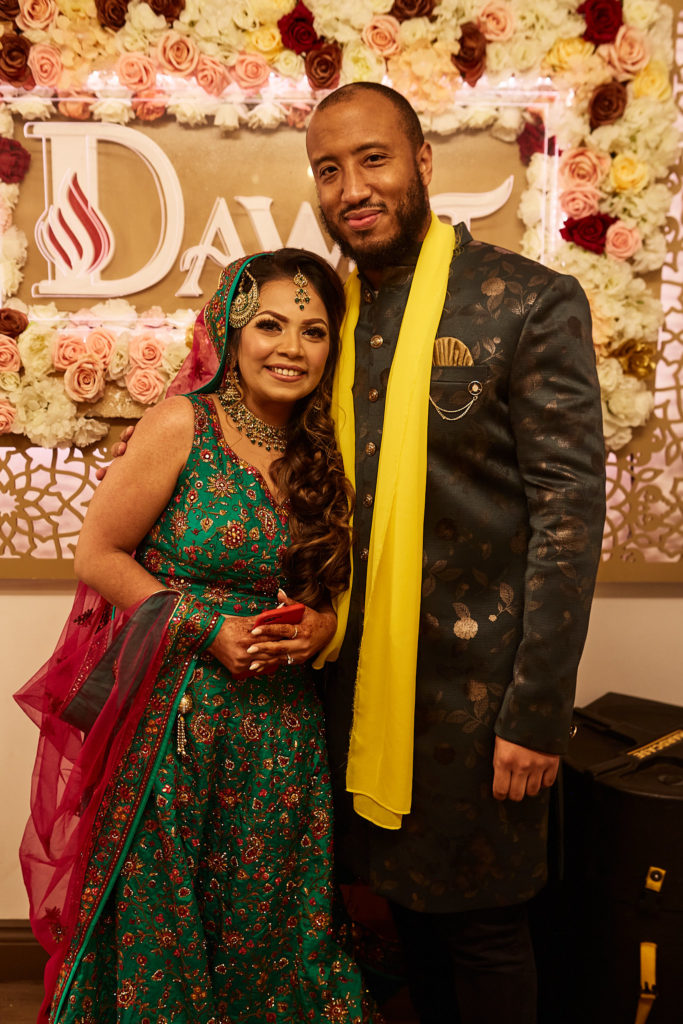 Bride in a green lehenga and groom in a yellow scarf in front of a flower wall and neon sign