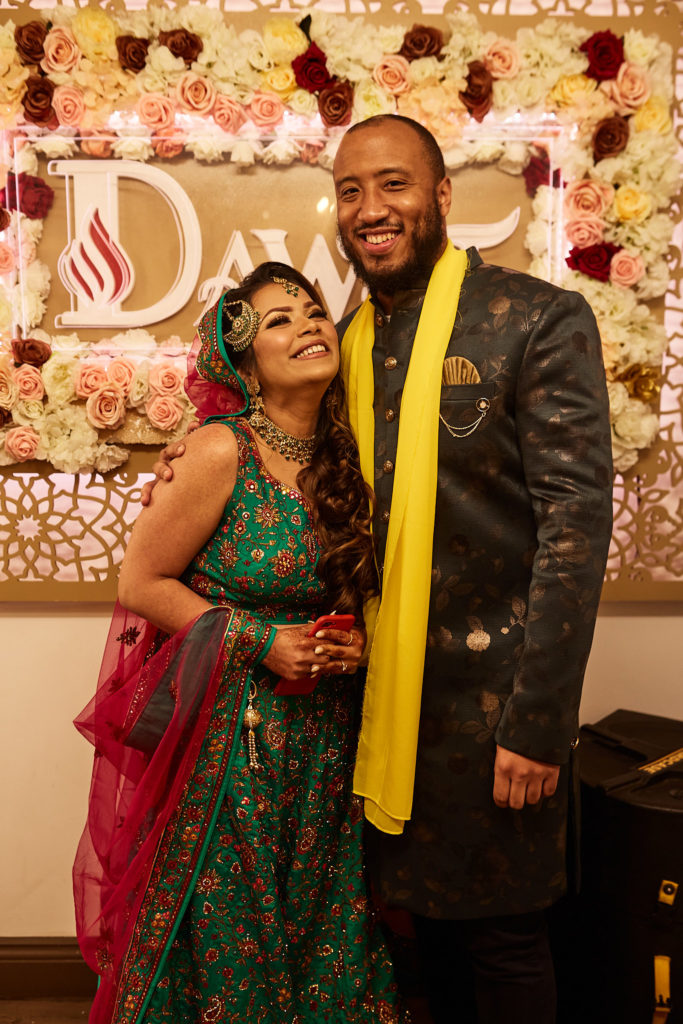 Bride looking up at the groom and laughing in front of the flower wall at their mehndi