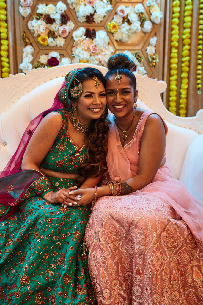 Bride in a green lehenga cuddling up to a friend in a pink lehenga on the stage sofa