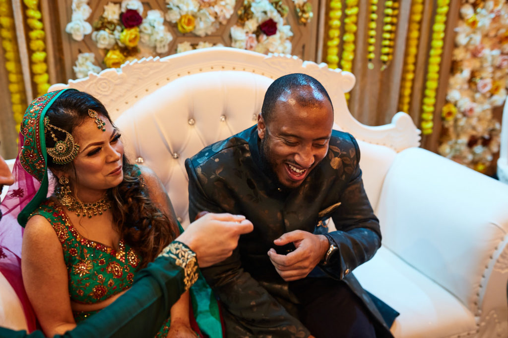 Bride laughing beside the groom on a white sofa in front of marigold garlands