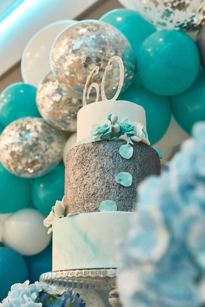 Three-tier 60th birthday cake in white, silver and turquoise among matching balloons