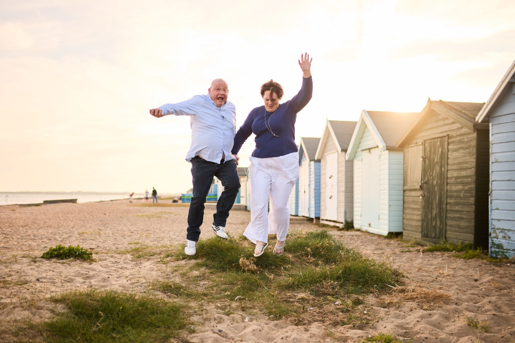 Engaged couple dancing along the beach beside a row of beach huts