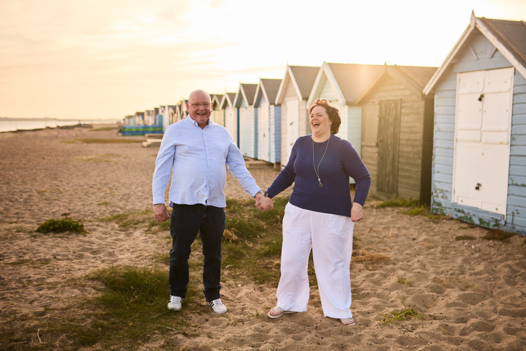 Engaged couple walking arm in arm past pastel beach huts on Mersea Island, Essex