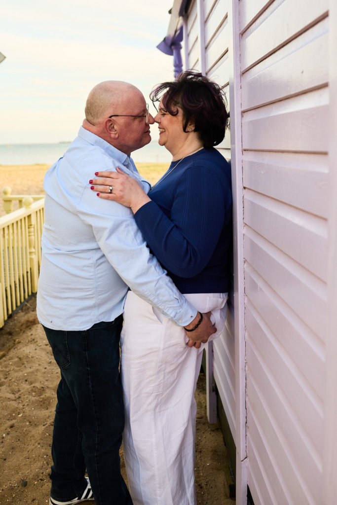 Engaged couple nose to nose beside a pink beach hut on Mersea Island
