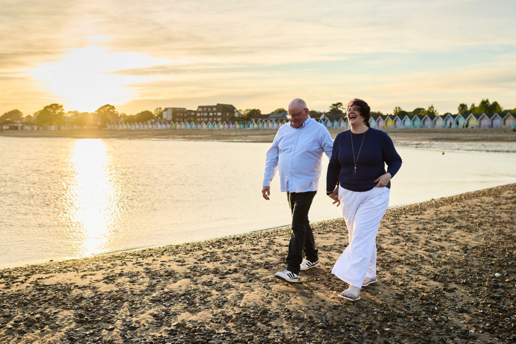 Engaged couple strolling along the shore as the sun sets over the water at Mersea Island