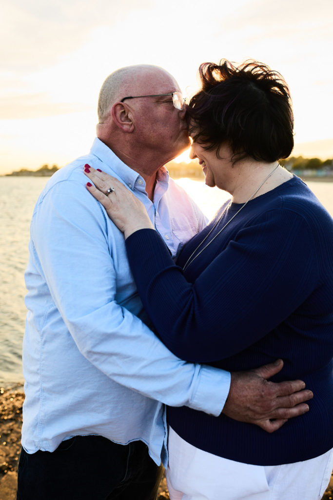 Man kissing his fiancée's forehead as they hug on the beach at sunset