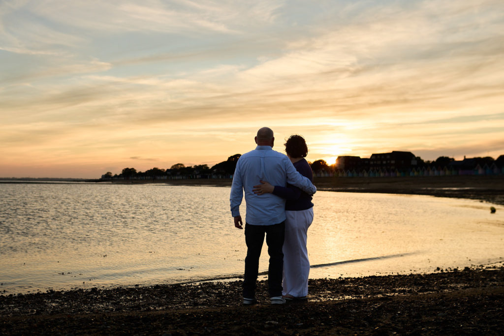 Engaged couple with their arms round each other watching the sunset over the water at Mersea Island
