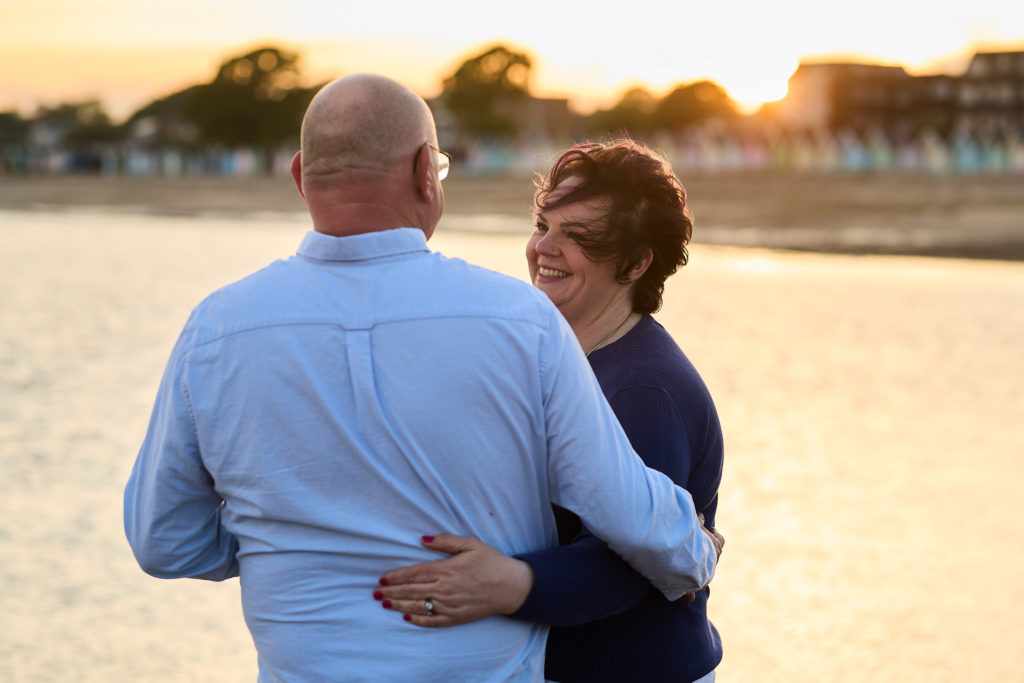 Woman smiling over her fiancé's shoulder as they stand by the water at sunset