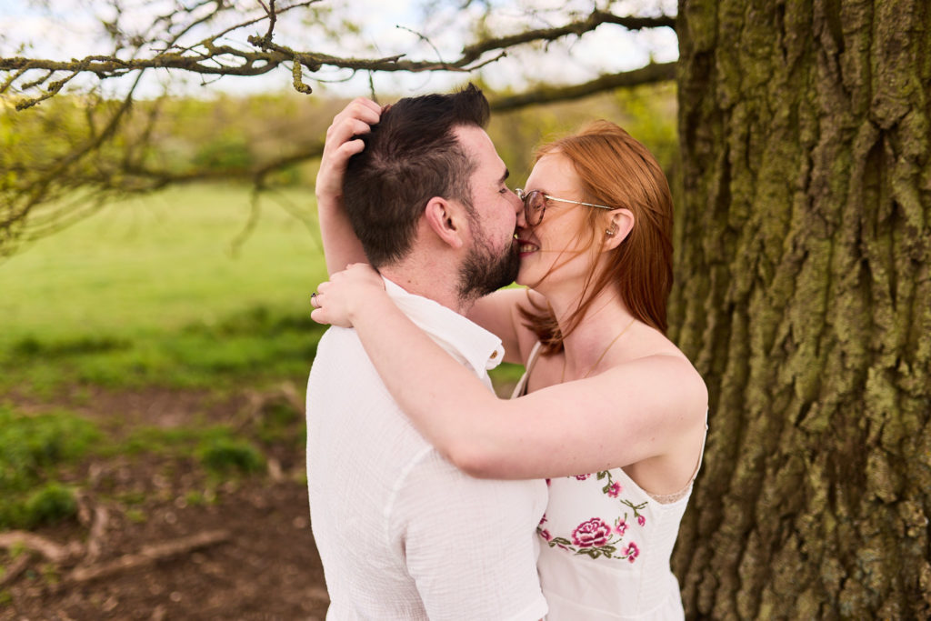 Engaged couple kissing beside a tree trunk in the Essex countryside