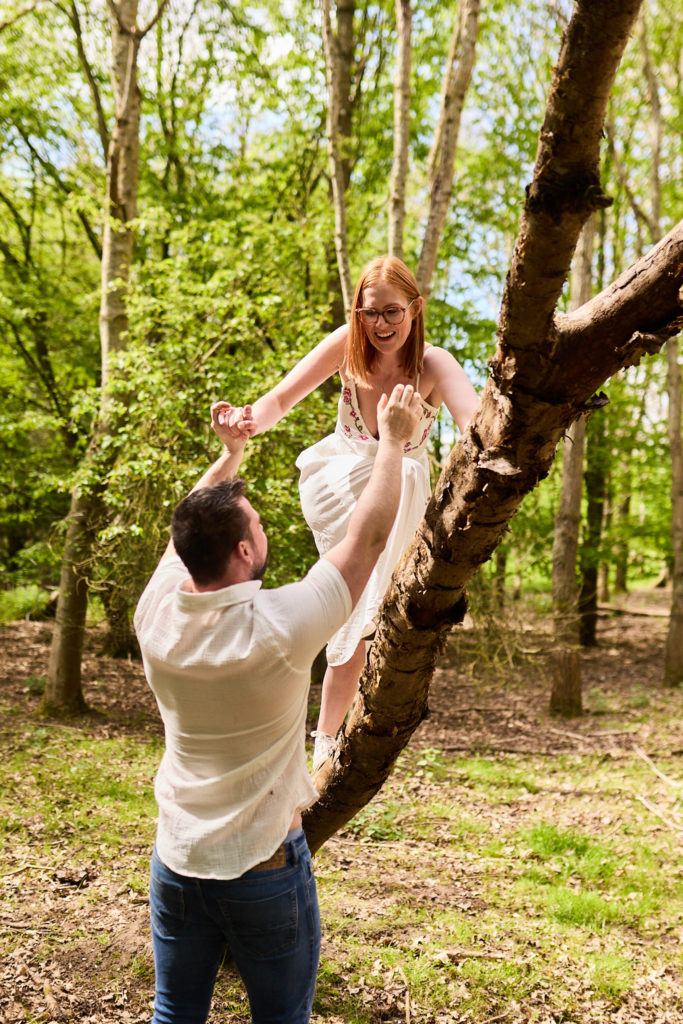 Woman perched in a tree reaching down to her fiancé during their woodland engagement shoot in Essex