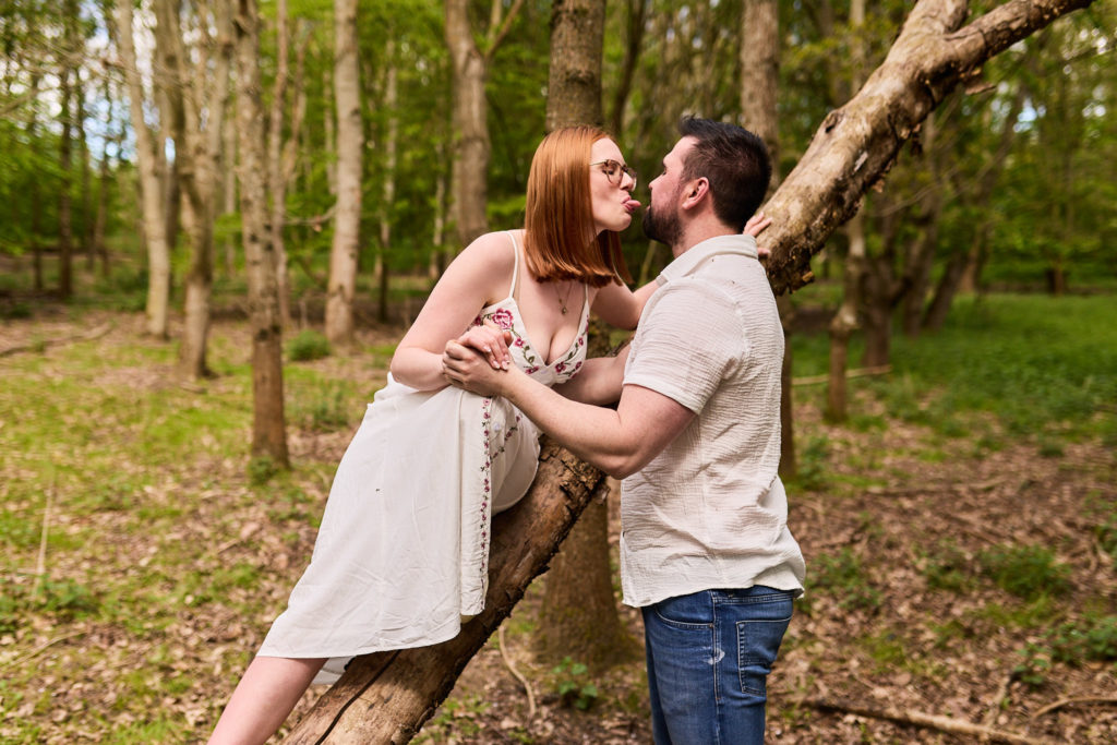 Woman sitting on a leaning tree trunk as her fiancé leans in for a kiss