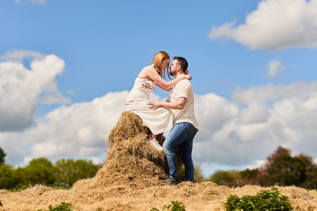Engaged couple kissing on top of a pile of hay under a blue sky