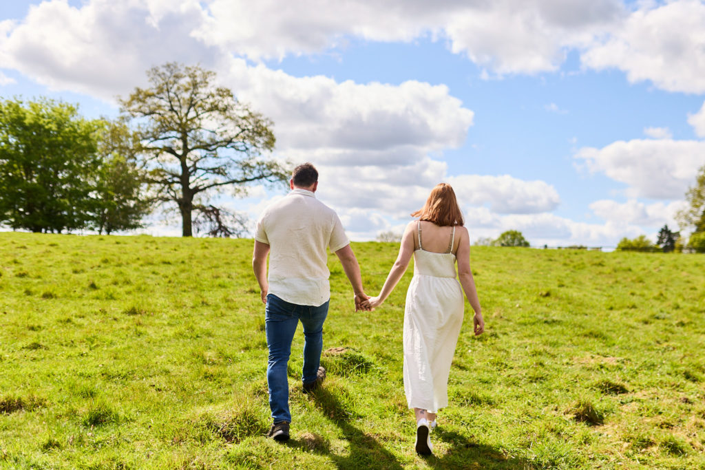 Engaged couple walking hand in hand across a field, seen from behind