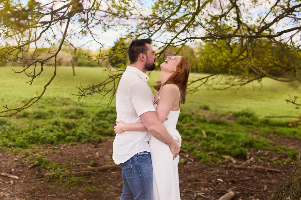 Man hugging his fiancée from behind as she turns to kiss him under a tree