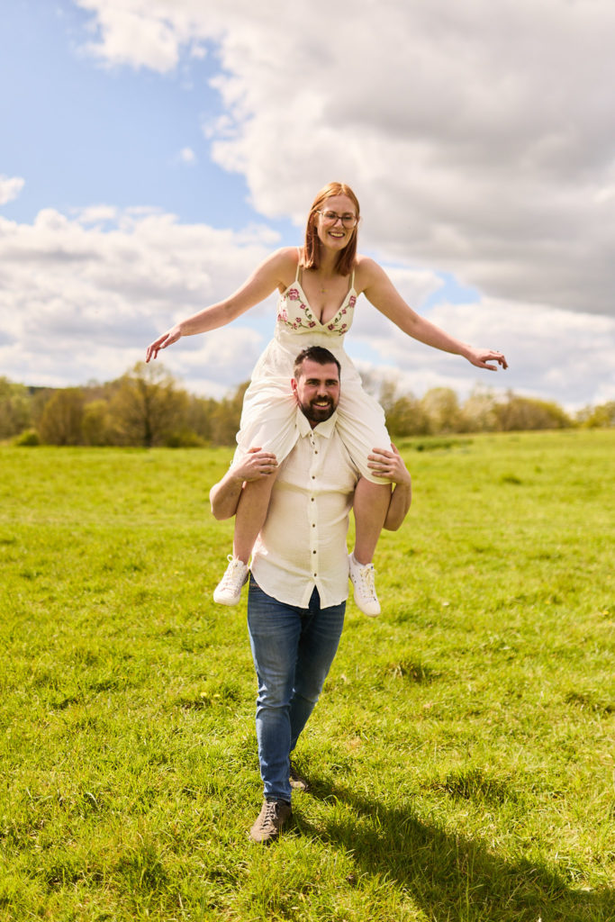 Woman riding on her fiancé's shoulders with her arms out across a sunny field