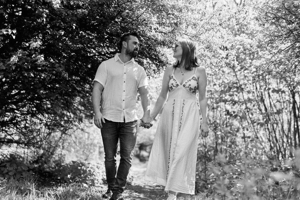 Black and white photo of an engaged couple walking hand in hand through the trees