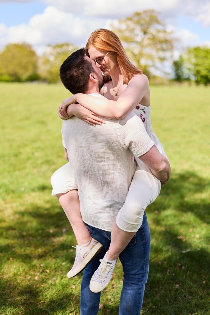 Man lifting his fiancée as they kiss in a sunny field