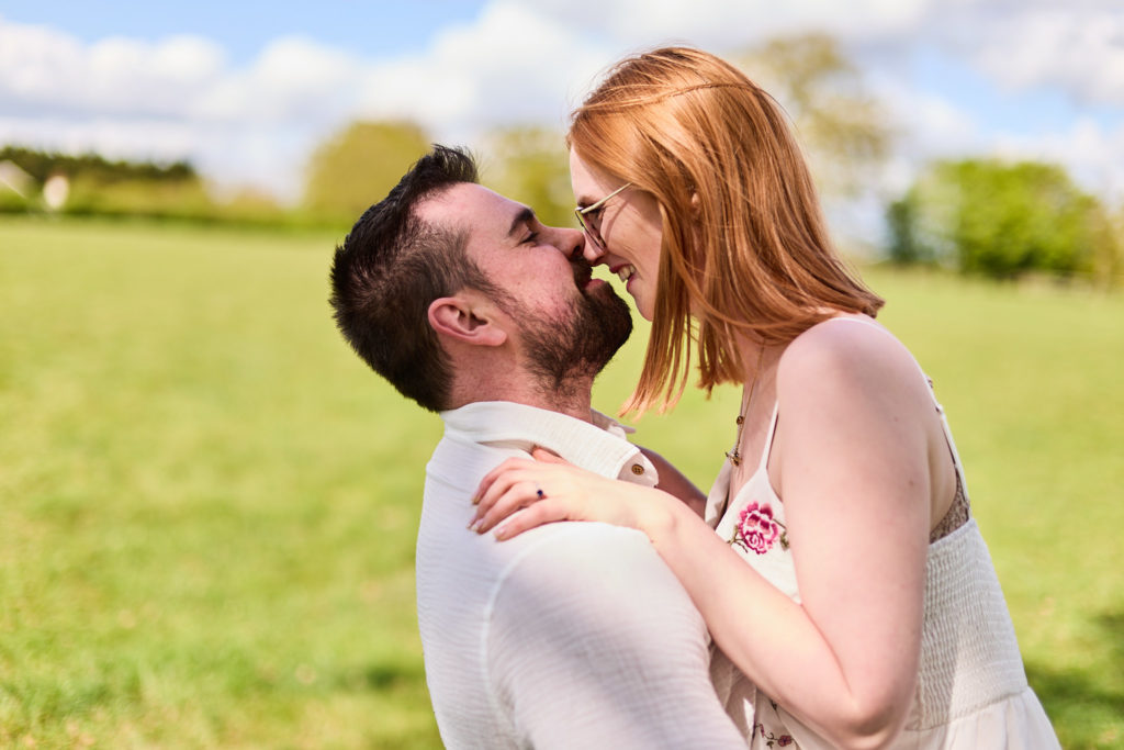 Engaged couple about to kiss in a field under a blue sky