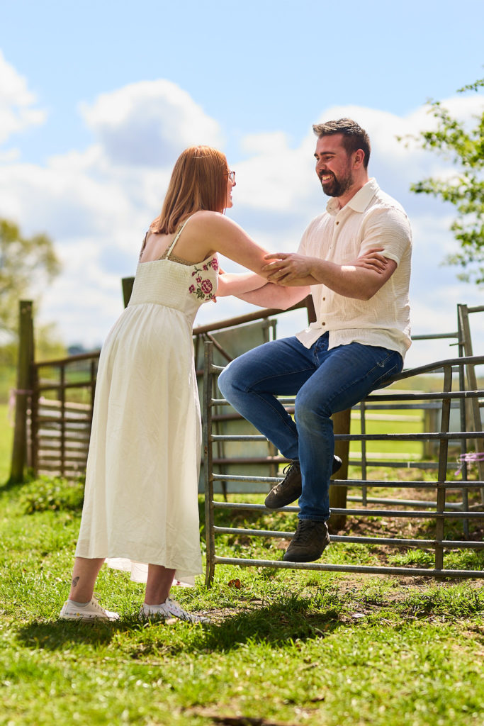 Woman leaning in to her fiancé as he sits on a field gate