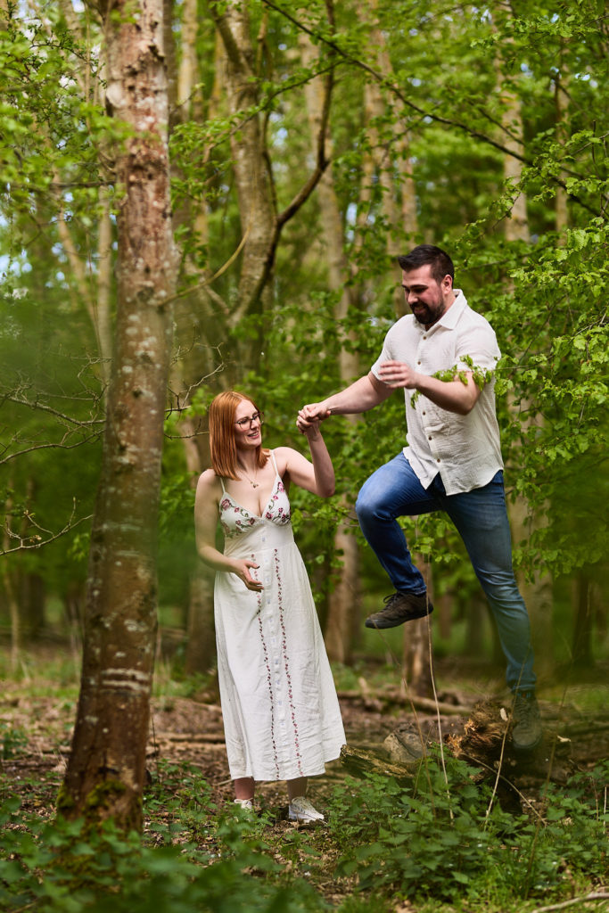 Man leaping in the air beside his laughing fiancée in the woods