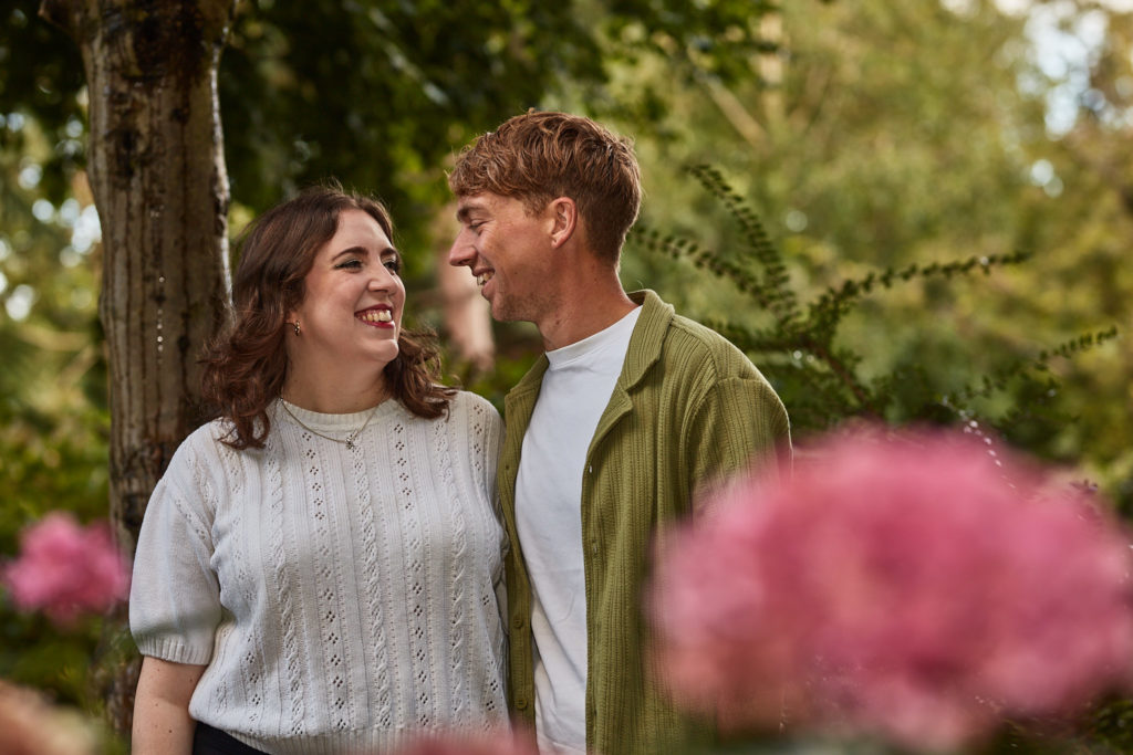 Engaged couple smiling at each other with pink flowers blurred in the foreground