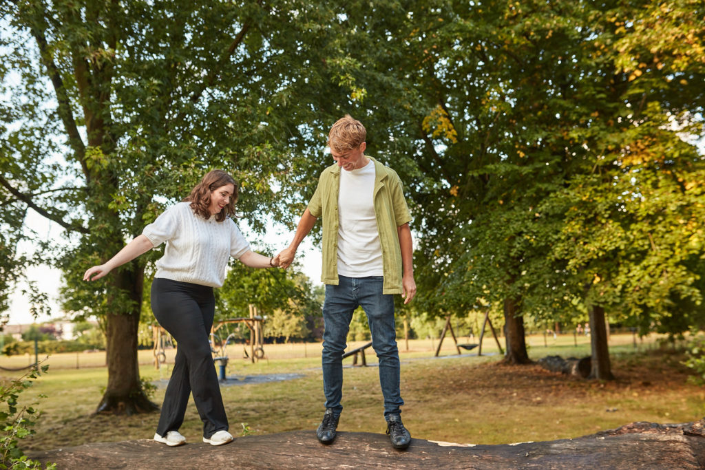 Engaged couple laughing with their arms stretched out for balance under park trees