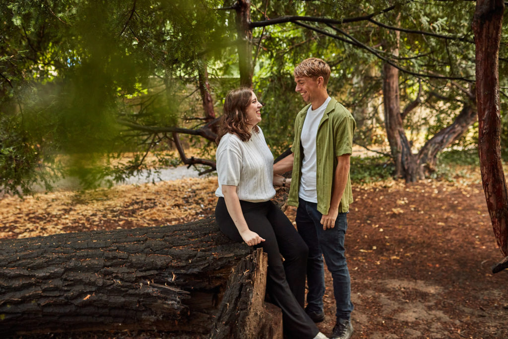 Woman leaning on a fallen log looking up at her fiancé, framed by green leaves