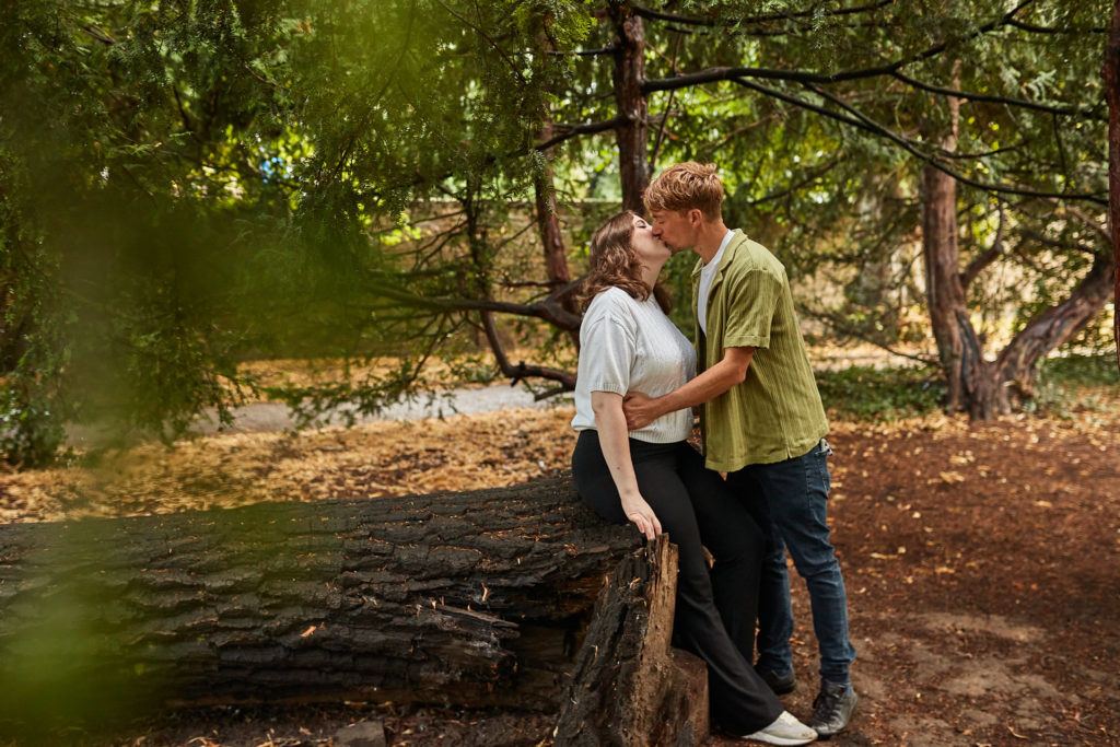 Engaged couple kissing beside a fallen log in woodland in Chelmsford