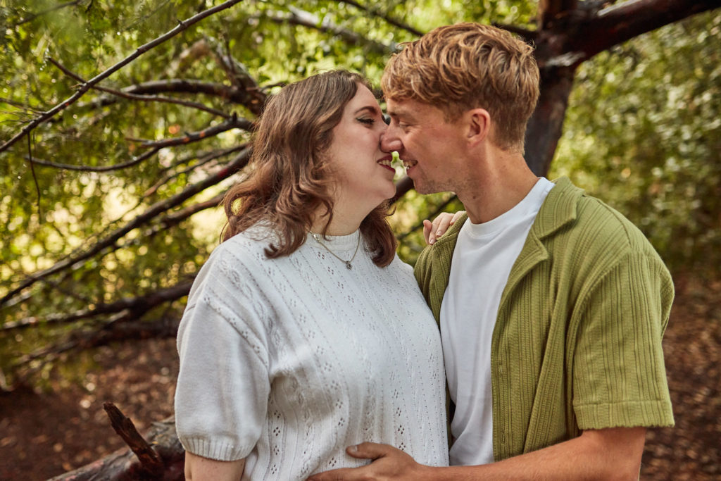 Man kissing his smiling fiancée on the cheek under pine branches