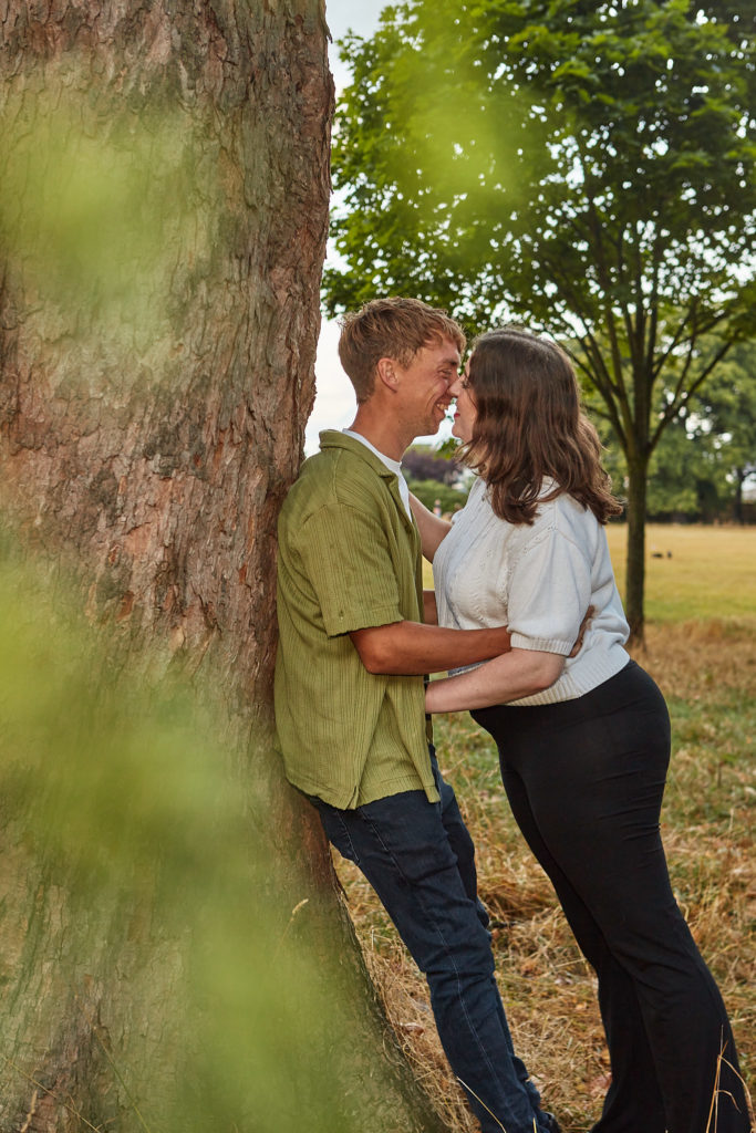 Engaged couple leaning face to face against a tree trunk