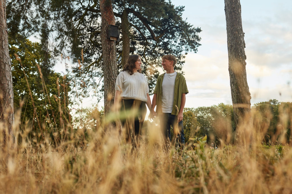Couple holding hands and smiling at each other between tall pine trees, seen through long golden grass on their engagement shoot