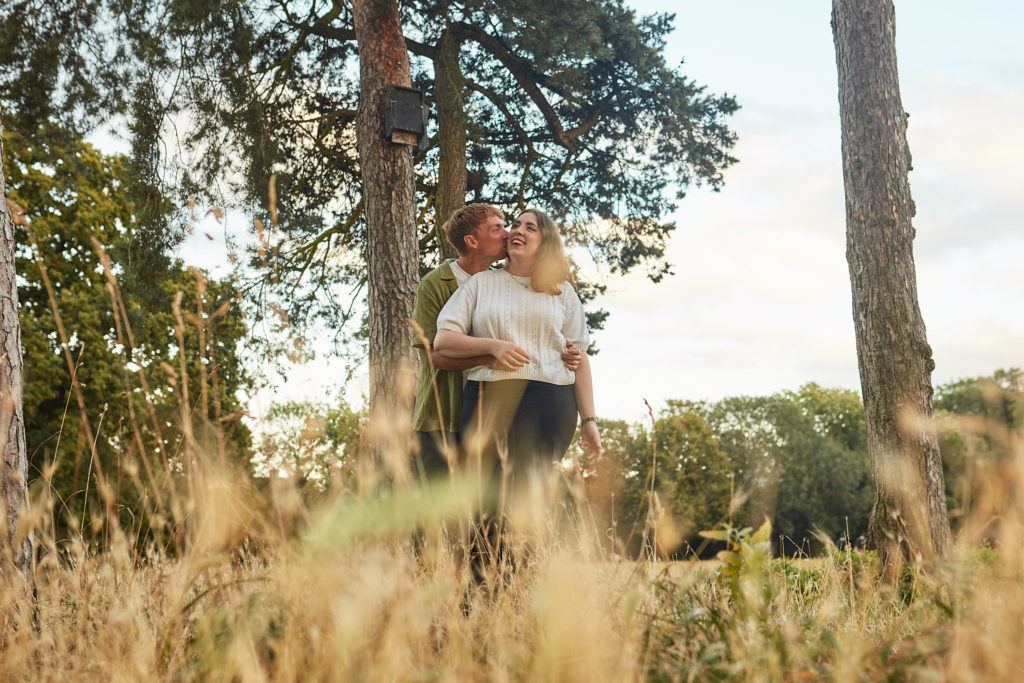 Engaged couple cuddling between tall pine trees, seen through long golden grass
