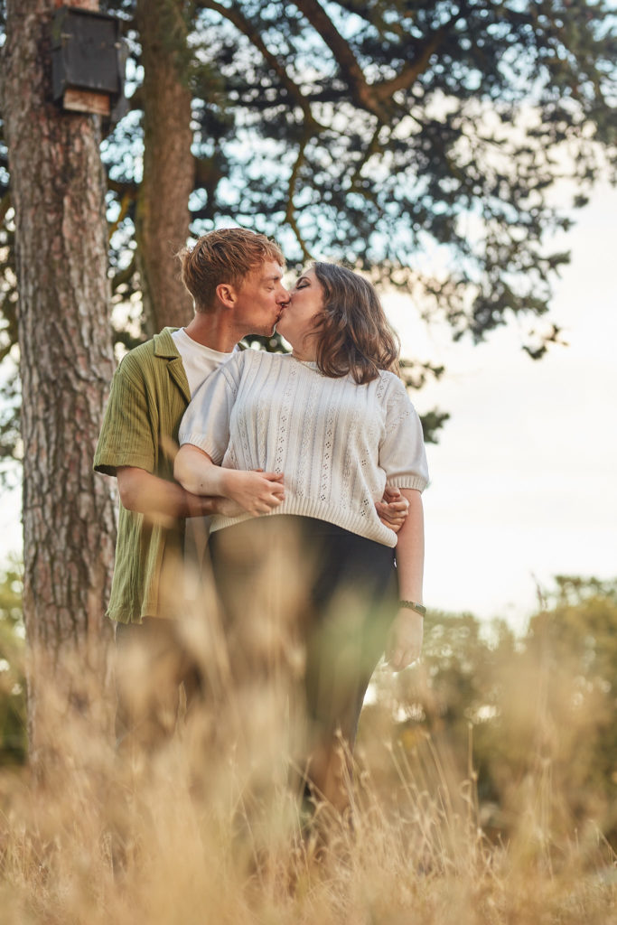 Man hugging his fiancée from behind and kissing her cheek in long grass beside a pine tree