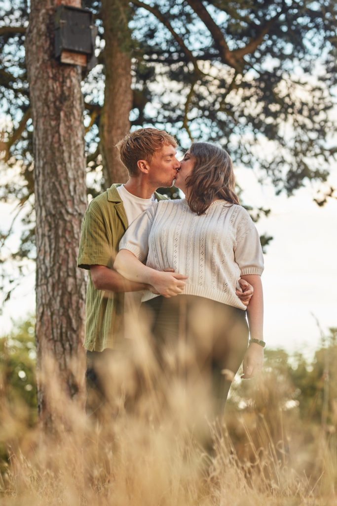 Man holding his fiancée from behind as they smile, standing in long grass under a pine tree