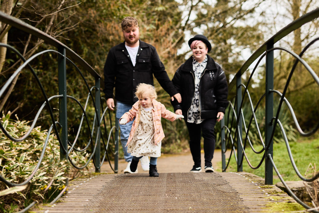 Couple walking hand in hand with their toddler across a footbridge in the park