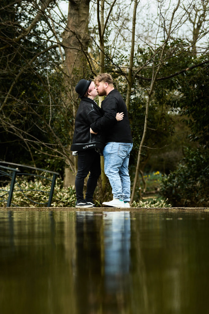 Engaged couple embracing beside a pond, reflected in the water