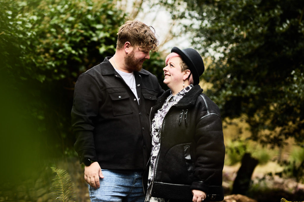 Man kissing his fiancée's cheek as she smiles, framed by green leaves