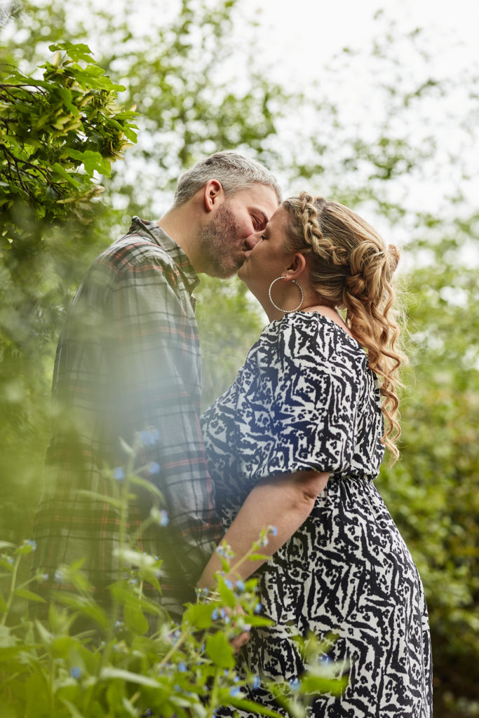 Engaged couple kissing in the park, framed by green foliage