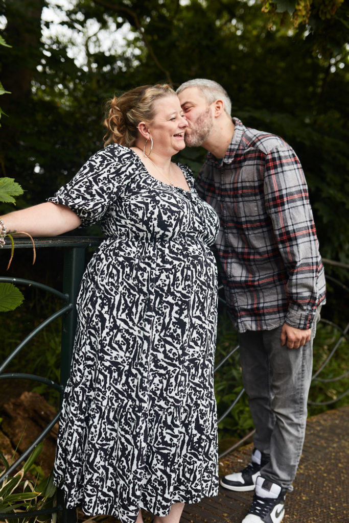 Man kissing his fiancée's cheek as she smiles on a footbridge